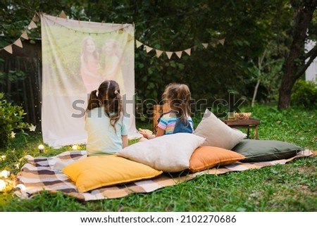 Backyard Family outdoor movie night with kids. Sisters spending time together and watching cimema at backyard. DIY Screen with film. Summer outdoor weekend activities with children. Open air cinema. Royalty-Free Stock Photo #2102270686
