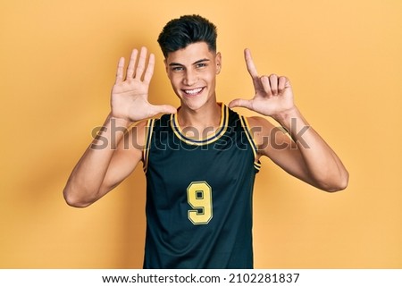 Young hispanic man wearing basketball uniform showing and pointing up with fingers number seven while smiling confident and happy. 