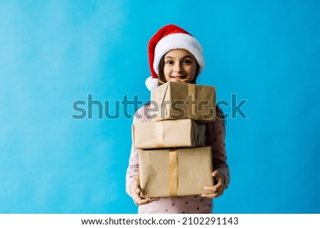 Happy little girl wearing christmas hat opening a gift box isolated on blue background.