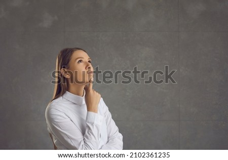 Portrait of a dreamy and pensive woman standing thoughtfully on a gray background. Young beautiful woman looks up and touches her chin, pondering a proposal or making a choice. Place for text.