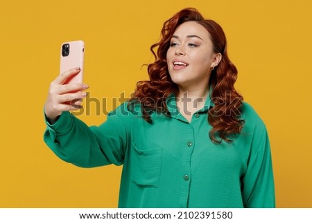 Bright happy vivid young ginger chubby overweight woman 20s wears green shirt doing selfie shot on mobile cell phone post photo on social network isolated on plain yellow background studio portrait
