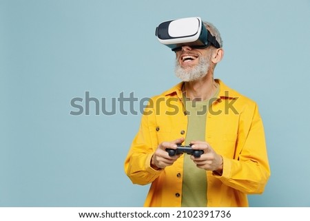 Elderly gray-haired mustache bearded man 50s wear yellow shirt hold in hand play pc game with joystick console watching in vr headset pc gadget isolated on plain pastel light blue background studio. Royalty-Free Stock Photo #2102391736