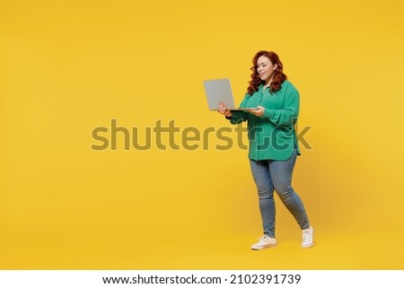 Full size body length bright happy vivid fancy young ginger chubby overweight woman 20s wears green shirt hold use work on laptop pc computer typing isolated on plain yellow background studio portrait