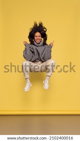 Fun, joy. Full-length portrait of young happy dark skinned girl in warm knitted sweater jumping isolated on yellow background. Concept of emotions, facial expression, youth, sales and aspiration.