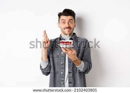 Birthday. Happy guy making wish with fingers crossed, holding bday cake with candle, standing on white background