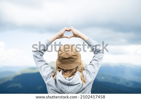 Womans making heart shape with her hands against amazing Carpathians mountain panorama. Traveler on mountain summit enjoying aerial view hands raised over clouds