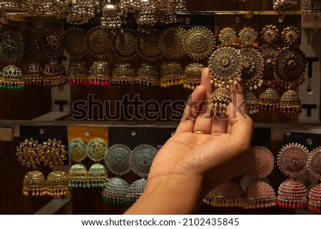 A hand of a woman selecting earrings on street market shop in Red fort, Delhi. Street shopping, bargain, ethnic, jewelry, cheap, expensive, women, girls, prices, sarojini nagar, chandni chowk concept. Royalty-Free Stock Photo #2102435845