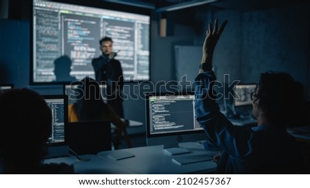 Teacher Giving Computer Science Lecture to Diverse Multiethnic Group of Female and Male Classmates in Dark College Room. Student Raises Hand and Answers Lecturer a Question About Software Engineering. Royalty-Free Stock Photo #2102457367
