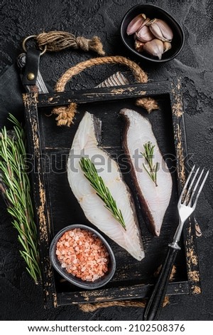 Raw halibut fish steak in wooden tray with herbs. Black background. Top view