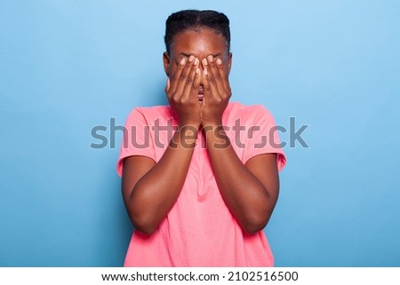 Portrait of scared stressed african american young woman covering face with her hands during photo shotting having social anxiety standing in studio with blue background. Terrified introvert model Royalty-Free Stock Photo #2102516500