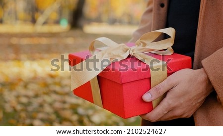 Boyfriend congratulates girlfriend unknown guy give gift unrecognizable woman in autumn city park young girl receive surprise red box with gold ribbon outdoors male hands holding present for beloved