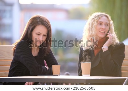 Female friends spending time at street cafe outdoors while one girl is talking happily on mobile phone and another waiting frustrated at offensive behaviour of her mate. Friendship problems concept Royalty-Free Stock Photo #2102565505