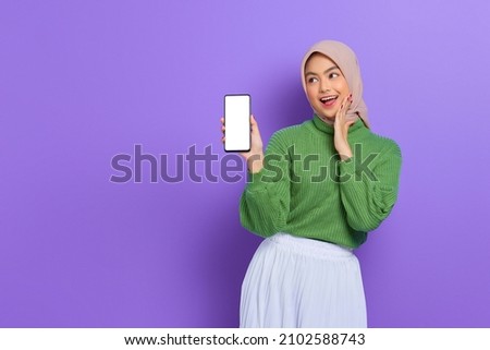 Smiling beautiful Asian woman in green sweater and hijab showing blank screen mobile phone isolated over purple background