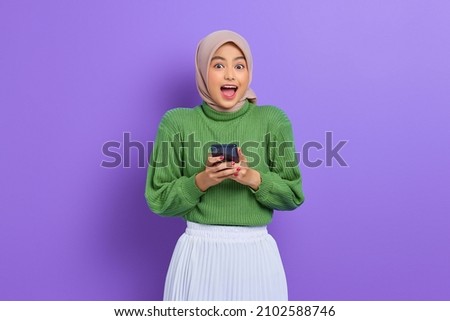 Excited beautiful Asian woman in green sweater and hijab using a mobile phone, received good news isolated over purple background Royalty-Free Stock Photo #2102588746