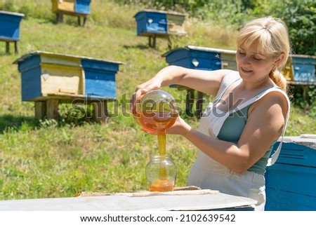 Harvest honey. woman beekeeper pours honey into a jar on the background of an apiary, beehives. Beautiful female girl fills a jar with honey on a sunny summer day in a green meadow