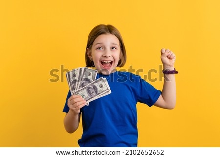 Excited Little Girl 8-10 Years Old in Basic Blue T-shirt on Yellow Background. Child Holding Bundle of Dollar Bills and Smiling. Children Financial Literacy Concept. Invest in Future