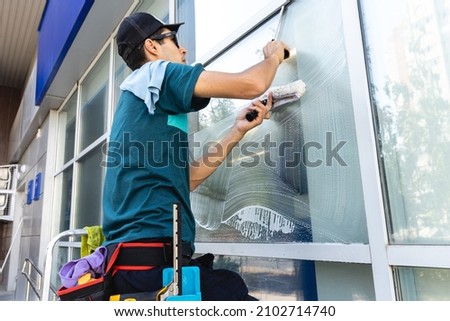 Male professional cleaning service worker in overalls cleans the windows and shop windows of a store with special equipment Royalty-Free Stock Photo #2102714740
