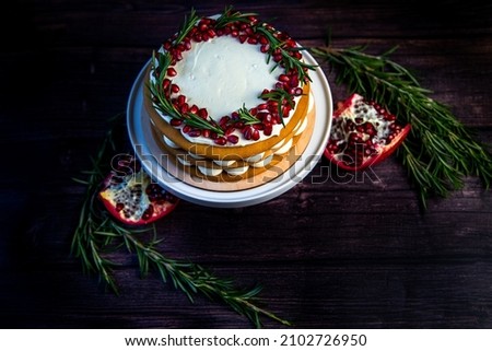 Cake with white cream and honey brown cakes lies on a white stand, decorated with pomegranate and rosemary, against a dark tree. High quality photo