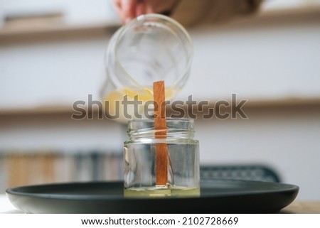 Close-up of unrecognizable female artisan pouring melted wax into tranparent glass jar with wick at home. Process of making handmade natural candle at workshop.