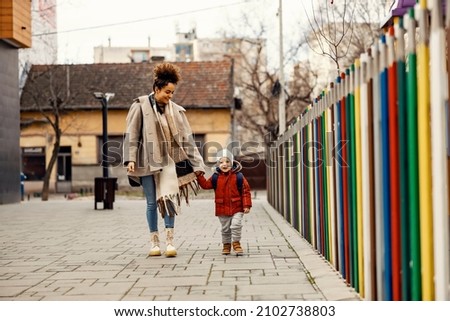 A young woman, nanny or caregiver, holding hands with a little boy with a schoolbag on his backs and walking outdoors. Royalty-Free Stock Photo #2102738803