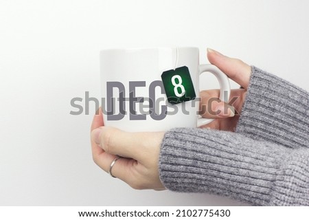 December 8th. Day 8 of month, Calendar date. Closeup of female hands in grey sweater holding cup of tea with month and calendar date on teabag label. Winter month, day of the year concept