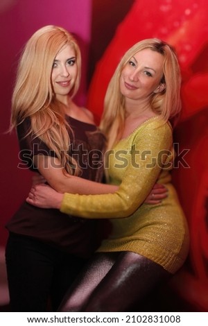 Two beautiful, fashionable blonde girls are smiling and hugging each other on a red background. Photo of young lesbian couple hugging each other while standing together at home. Lgbt photo