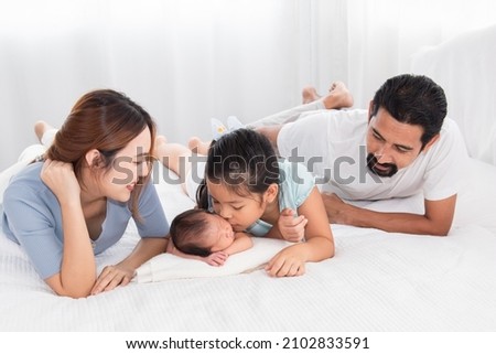 Asian parents and newborn with sister spend time together in bedroom. Sister kiss Adorable infant while baby sleeping among happy family, mother and father lying in bed with two daughters.