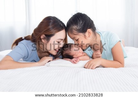 Beautiful Asian mother and daughter spend time with newborn in bed, two woman kiss adorable infant with love and care. Toddler baby sleeping in bedroom with family.