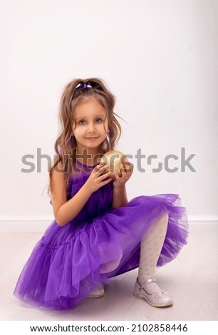 A girl in a purple dress and with beautiful curls holds a golden Christmas ball in her hand on a white background