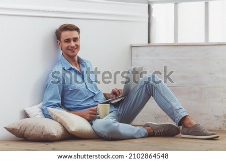 Young handsome man in the blue shirt and jeans, with a cup in hand, using laptop, sitting on cushions on the floor, next are graphics, sitting on the floor at home