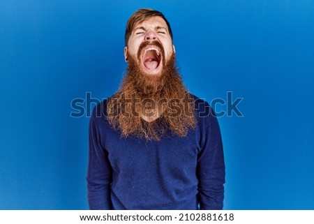 Redhead man with long beard wearing casual blue sweater over blue background angry and mad screaming frustrated and furious, shouting with anger. rage and aggressive concept. 