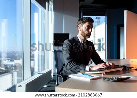 Focused broker working at computer in marvelous office by the window, male businessman in business suit