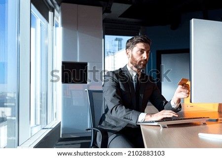 Serious businessman shopping online sitting in the office, working at the computer, man holding a credit card, sitting at a table by the window