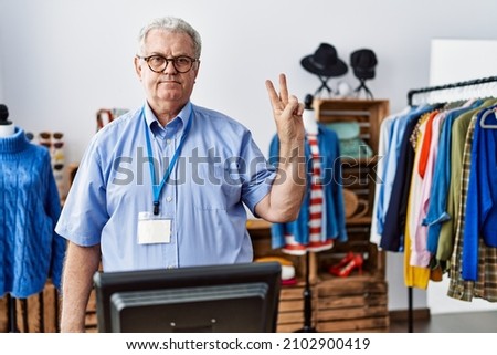 Senior man with grey hair working as manager at retail boutique showing and pointing up with fingers number two while smiling confident and happy. 
