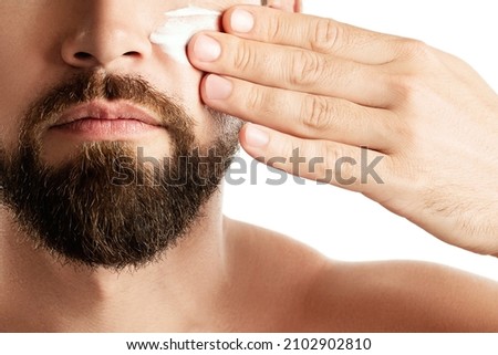Men's beauty - Young man is applying moisturizing and anti aging cream on his face against white background Royalty-Free Stock Photo #2102902810
