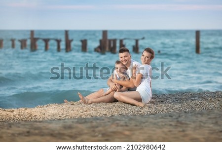 a happy family of a man, a woman and their baby boy are sitting on the seashore in the evening
