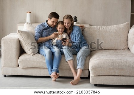 Full length joyful loving young couple parents and cute small kid daughter watching funny video in social network on cellphone, using software applications, web surfing information or shopping online.