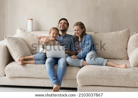 Emotional joyful laughing bonding family young parents and little preteen child daughter watching funny film or cartoons online on computer, resting on cozy sofa, modern technology gadget use.