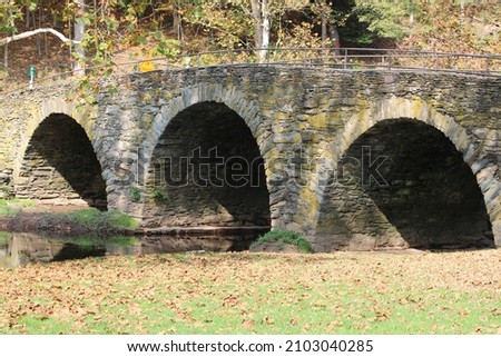 The historical Stone Arch Bridge in the Catskills. The stone footbridge has three arches, is full of falling, Autumn leaves, and green grass. The water below is reflecting the image of the bridge.