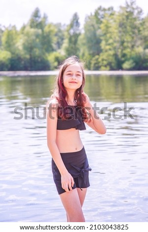 Children and rest. Teenage girl by the river in a black swimsuit and black shorts in summer.