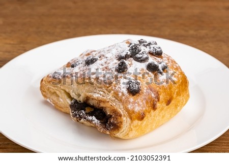 Single piece of Danish Pastry filled with chocolate cream topping with chocolate chips and icing sugar powder in white ceramic dish on wooden table.