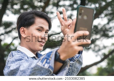 A young man poses for a selfie using his phone. A very active social media user. Standing outside a park.