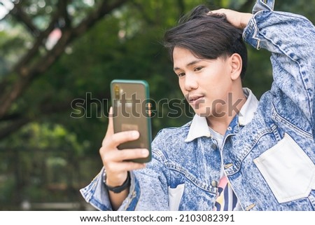 A young man checks his appearance using the front camera of his cellphone. Standing outside a park.