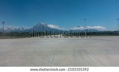 Marking lines and tire tracks are visible on the concrete runway of the airfield. Snow-capped conical volcanoes against a background of blue sky and clouds. Copy space. Petropavlovsk-Kamchatsky Royalty-Free Stock Photo #2103105392