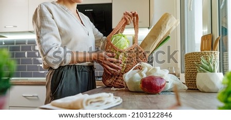 Young woman holding reusable mesh bag with fruits, vegetables and bread in the kitchen. Zero waste and sustainable packaging concept. Horizontal panoramic banner. Royalty-Free Stock Photo #2103122846