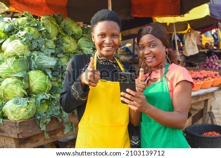 Two happy African business women or female traders wearing colorful aprons while standing at a vegetable stall in a market place with a smart phone with them Royalty-Free Stock Photo #2103139712