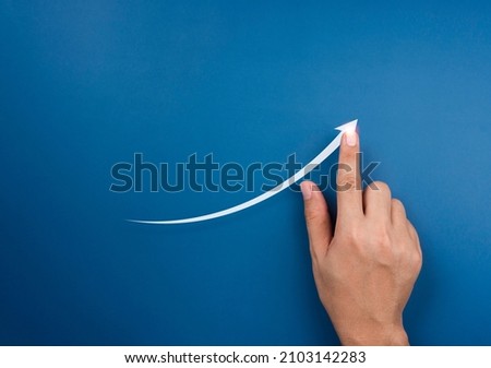 Hand draws a rising arrow on blue background. Finger pointing on the shining arrowhead, minimal style. Business growth and success concept. Increase business opportunities. Royalty-Free Stock Photo #2103142283