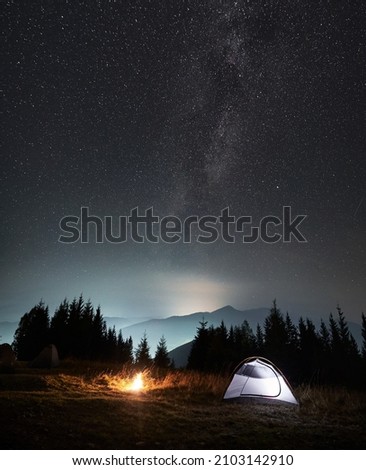 Magnificent view of night starry sky and Milky way over grassy hill with illuminated camp tent and campfire. Concept of hiking, night camping and astronomy. Royalty-Free Stock Photo #2103142910