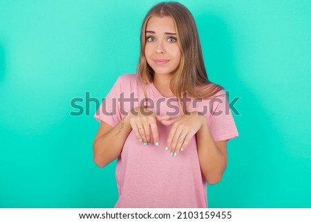 Young caucasian girl wearing pink T-shirt isolated over green background makes bunny paws and looks with innocent expression plays with her little kid