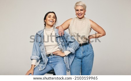Best friends wearing denim jeans in a studio. Two happy women smiling at the camera cheerfully. Fashionable female friends posing together against a grey background. Royalty-Free Stock Photo #2103159965
